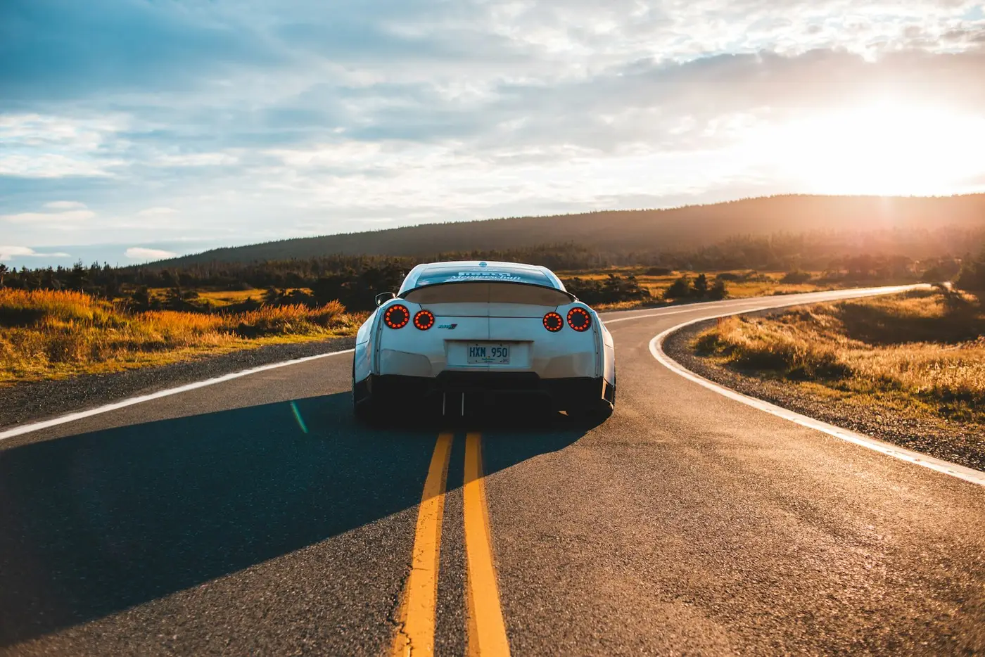 מכונית ספורט כסופה מסוג ניסאן GT-R מתרחקת בכביש מתפתל עם קו צהוב כפול בשקיעה, גבעות ברקע