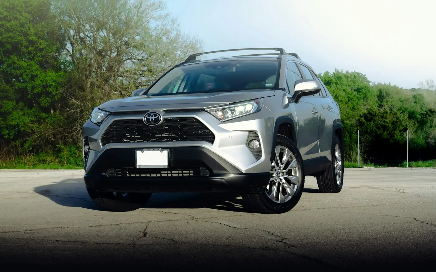 רכב פנאי טויוטה RAV4 אפור, מבט מלפנים, במגרש חניה ריק עם עצים ברקע